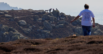 Walkers in Cornwall This landscape photograph, taken in the late morning during early spring, captures walkers exploring the rugged coast of Cornwall in the United Kingdom. The image showcases the natural beauty of the area, with rocky outcrops and coastal vegetation stretching towards the horizon, where the sea meets the misty sky. Prominent in the foreground is a person dressed in casual attire, leading a dog along the undulating terrain, while other walkers and another dog can be seen further in the distance, highlighting the region's appeal for outdoor recreation and nature enthusiasts. The composition and lighting emphasize the unique characteristics of Cornwall’s coastal environment, making it a representative scene of nature along the United Kingdom's southwest shoreline.